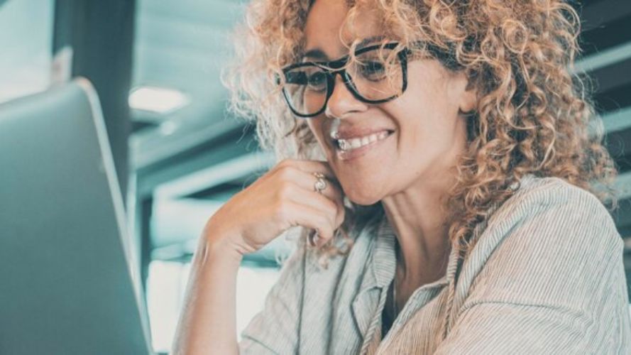 Smiling lady with curly hair and glasses reads blog on laptop